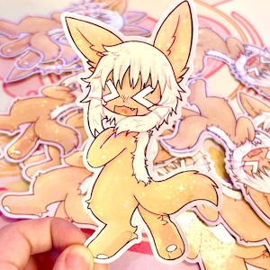 May include: A sticker featuring a cartoon character with large ears, white hair, and a scarf. The character has a surprised expression with crossed-out eyes and an open mouth. The sticker has a glittery finish and is held by a hand, with other similar stickers in the background.