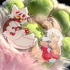 May include: Two clear acrylic charms featuring anime-style characters. One charm has a white and brown character with red accents. The other charm has a white and tan character with pink and red accents. The charms are on a fluffy pink and green background.