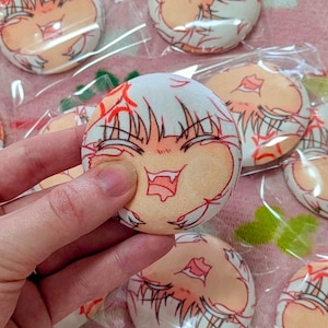 May include: A collection of round, plush items featuring a cartoon face design. The faces have white hair, orange skin, and red accents. Some are held in a hand, while others are packaged in clear plastic.