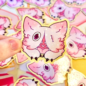 May include: A pink and white sticker featuring a cartoon creature with large eyes, claws, and a heart-shaped mouth. The sticker has a holographic, glittery finish and a yellow border. Other similar stickers are visible in the background.