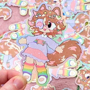 May include: A holographic sticker featuring a cartoon fox-like character with brown fur, wearing a purple top, pink skirt, and rainbow socks. The character has a patch over one eye and a fluffy tail. Other stickers are in the background.