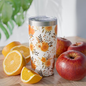 May include: A white stainless steel tumbler with a floral pattern featuring orange, yellow, and brown flowers. The tumbler is sitting on a wooden surface with oranges and red apples.