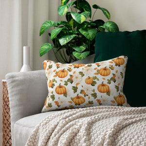 May include: A white throw pillow with a pattern of orange pumpkins and white flowers on a white background. The pillow is sitting on a white couch with a green throw blanket.