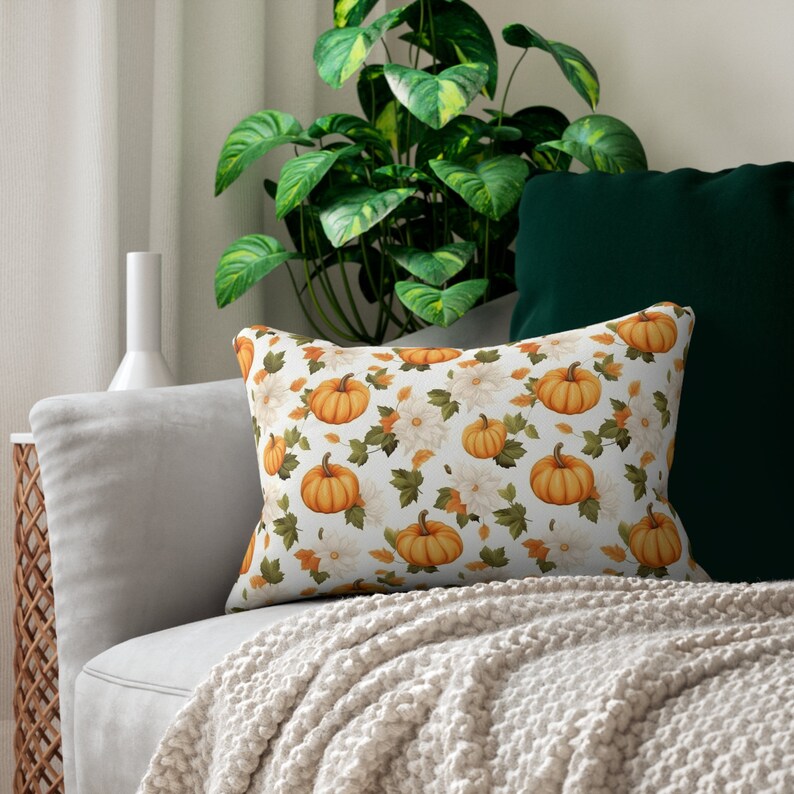 May include: A white throw pillow with a pattern of orange pumpkins and white flowers on a white background. The pillow is sitting on a white couch with a green throw blanket.