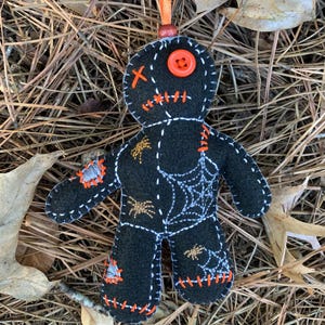May include: A black felt doll ornament with orange stitching and accents. The doll features a button eye, spiderweb and spider embroidery, and an orange ribbon for hanging. The doll is lying on a bed of pine needles and leaves.