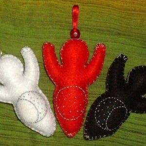 May include: Three felt ornaments in white, red, and black, each with a stitched design and a ribbon loop for hanging. The ornaments are in the shape of a person with arms outstretched.