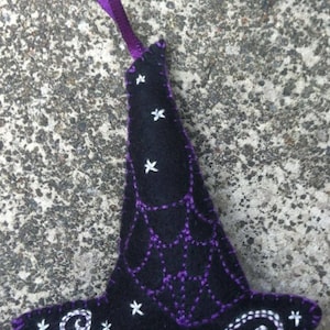 May include: A black felt witch hat ornament with purple stitching and a purple ribbon. The hat features a spiderweb design and white star accents, perfect for Halloween decorations.