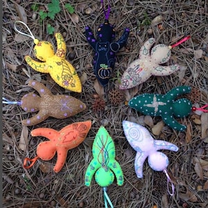 May include: A collection of handmade felt ornaments in various colors, including yellow, brown, orange, green, and purple. Each ornament has a unique design, such as a cat, a pumpkin, or a rocket ship, and is adorned with embroidered details and hanging loops.