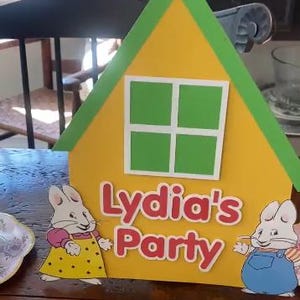 May include: A yellow house-shaped party decoration with a green roof and window. The text "Lydia's Party" is in red. Two cartoon bunny figures are on either side of the house.