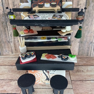 May include: A miniature sushi restaurant with a black counter, two black stools, and a sign that says "Let's Roll". The restaurant is decorated with Japanese-inspired patterns and features a variety of sushi rolls and other Japanese food.