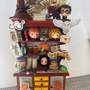 May include: A miniature, red wooden cabinet with multiple shelves, drawers, and decorative elements. The cabinet is adorned with various Harry Potter-themed figurines, books, and accessories, including a Sorting Hat, glasses, and a Hogwarts scarf. The cabinet has a Gryffindor crest on the door.