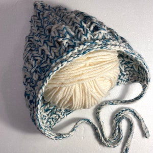 Puede incluir: Un gorro de bebé de punto blanco y azul con una bola de lana blanca en su interior. El gorro tiene una larga cinta que se puede atar debajo de la barbilla.