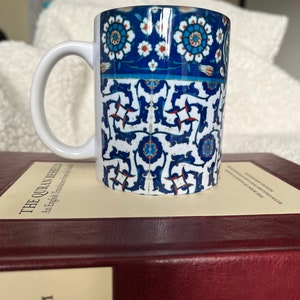 Könnte beinhalten: Eine weiße Keramiktasse mit einem blauen und weißen Blumenmuster. Die Tasse steht auf einem roten ledergebundenen Buch mit dem Titel "Der Koran"