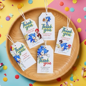 以下が含まれることがあります： ソニック・ザ・ヘッジホッグのキャラクターが描かれた、青と赤の「Thank you」の文字が入った白いギフトタグ。白いリボンが付いており、木製のトレイに並べられています。「For making my day unforgettable」の文字も含まれています。