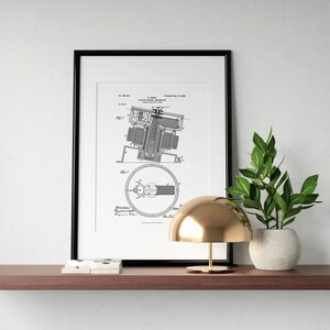 May include: A black framed print of a vintage patent drawing for a "Coupling for Electric Motors" hangs on a wall above a wooden shelf. A gold table lamp with a dome-shaped shade sits on the shelf next to a potted green plant.