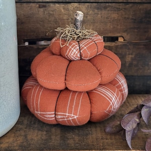 May include: A stacked fabric pumpkin decoration consisting of two rounded, stuffed sections in a burnt orange colour. The top section features a white plaid pattern, while the bottom section has a white plaid pattern. A small twig serves as the stem.