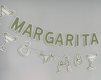 May include: A decorative banner featuring the text MARGARITA VEIL in sage green block letters. The banner is strung on a thin cord and includes alternating cutouts of margarita glasses, diamond rings, bikini tops, and lime slices in green.