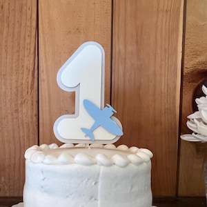 May include: A white frosted cake with a decorative border, topped with a number one cake topper and a blue airplane. The cake sits on a white scalloped edge cake board. The background is a wooden wall.