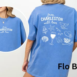 May include: A periwinkle blue t-shirt featuring white screen-printed graphics. The front displays the text "from CHARLESTON with love". The back features the same text above illustrations of a bikini, starfish, whale tail, wine glass, seagulls, sandals, sun hat, and coral, with "olivia's bachelorette" text.