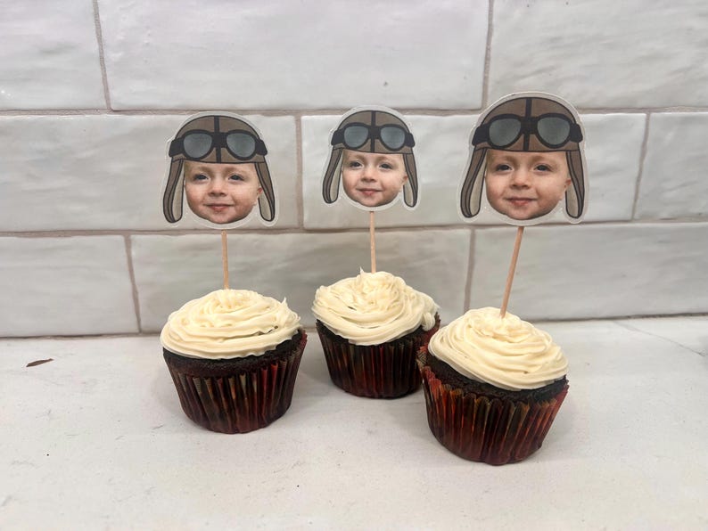 May include: Three chocolate cupcakes with vanilla frosting topped with personalized edible images of a child wearing a pilot hat and goggles.