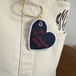 May include: Cream-colored tote bag with a gold-colored ring and a heart-shaped tag. The tag is navy blue with white trim and red text that reads "From Scottsdale Queen Cake". A brown monogram is embroidered on the bag.