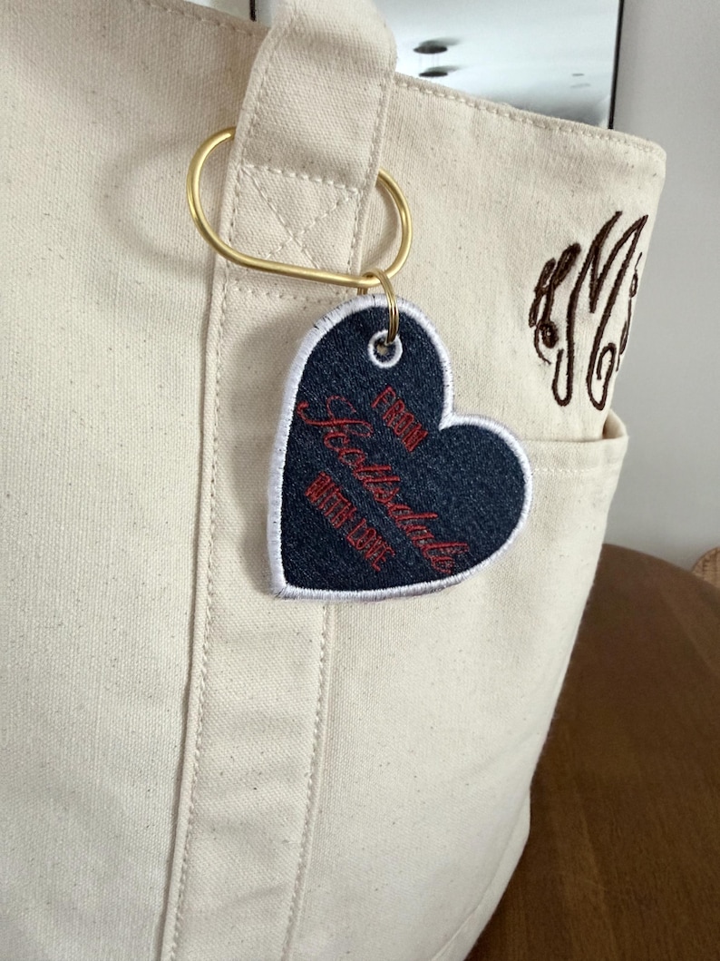 May include: Cream-colored tote bag with a gold-colored ring and a heart-shaped tag. The tag is navy blue with white trim and red text that reads "From Scottsdale Queen Cake". A brown monogram is embroidered on the bag.