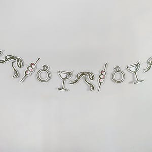 Op de afbeelding: Een decoratieve slinger met een herhalende reeks papieren uitsparingen opgehangen aan een dun touwtje. De illustraties bevatten diamanten ringen, martiniglazen, gestrikte linten en prikkers met drie olijven, getekend in zwarte inkt.