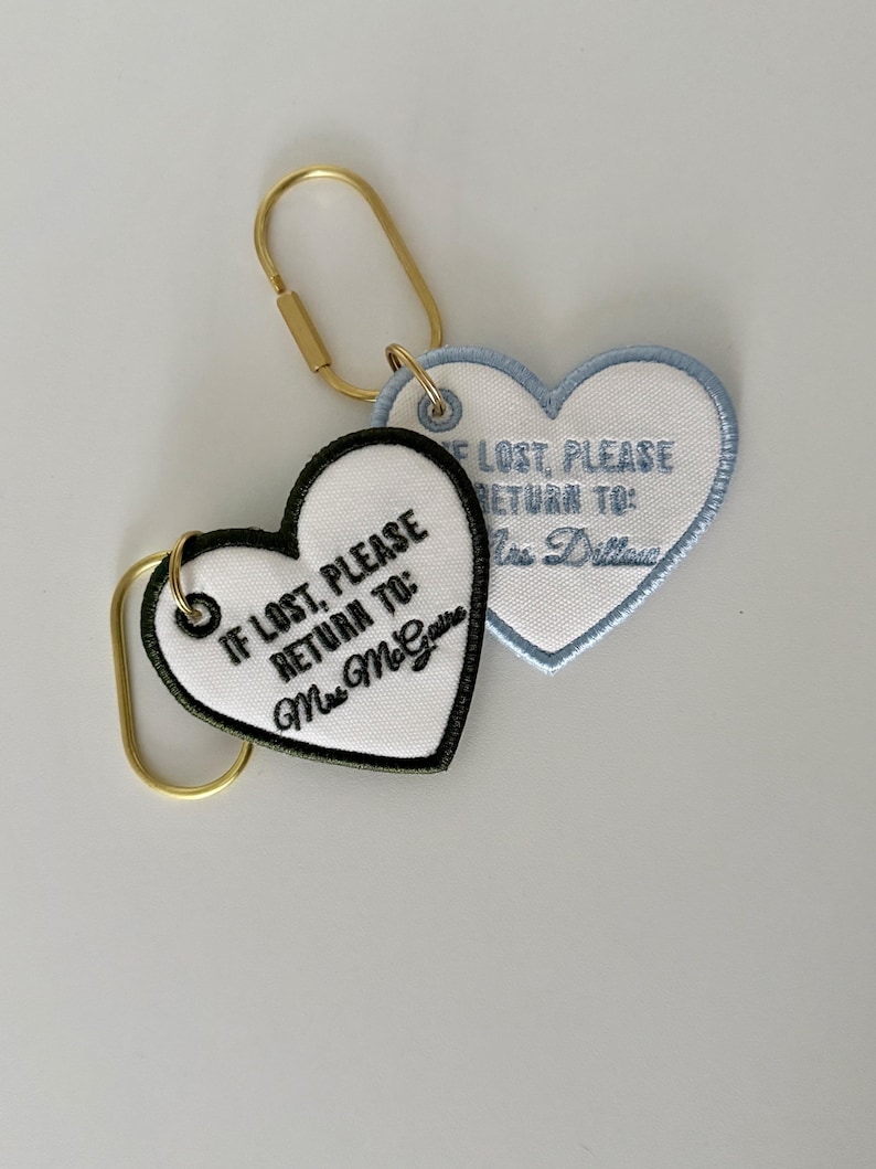 Personalized heart-shaped embroidered bag tag charms reading “If Lost, Please Return To,” featuring custom names, white fabric, colored stitching, and gold keyrings.