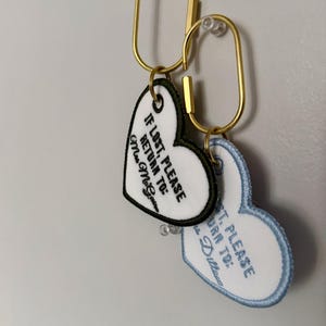 May include: Two heart-shaped keychains with gold-colored clasps. One keychain has a white background with black text that reads "IF LOST, PLEASE RETURN TO: Mrs. McGovern". The other keychain has a light blue border and text.