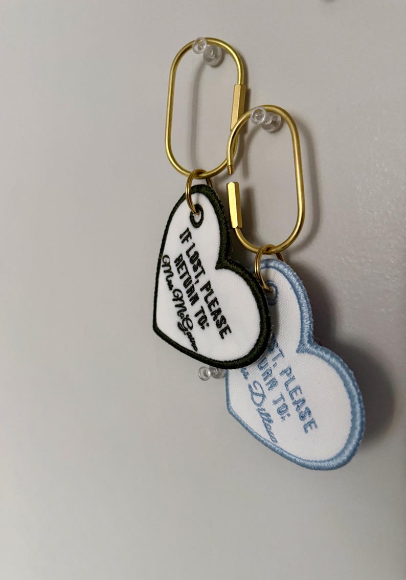May include: Two heart-shaped keychains with gold-colored clasps. One keychain has a white background with black text that reads "IF LOST, PLEASE RETURN TO: Mrs. McGovern". The other keychain has a light blue border and text.