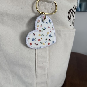 May include: A cream-colored tote bag with a gold-colored ring holding a heart-shaped keychain. The keychain has a white background with a floral pattern in blue, pink, and yellow. The bag has a pocket with embroidered initials.
