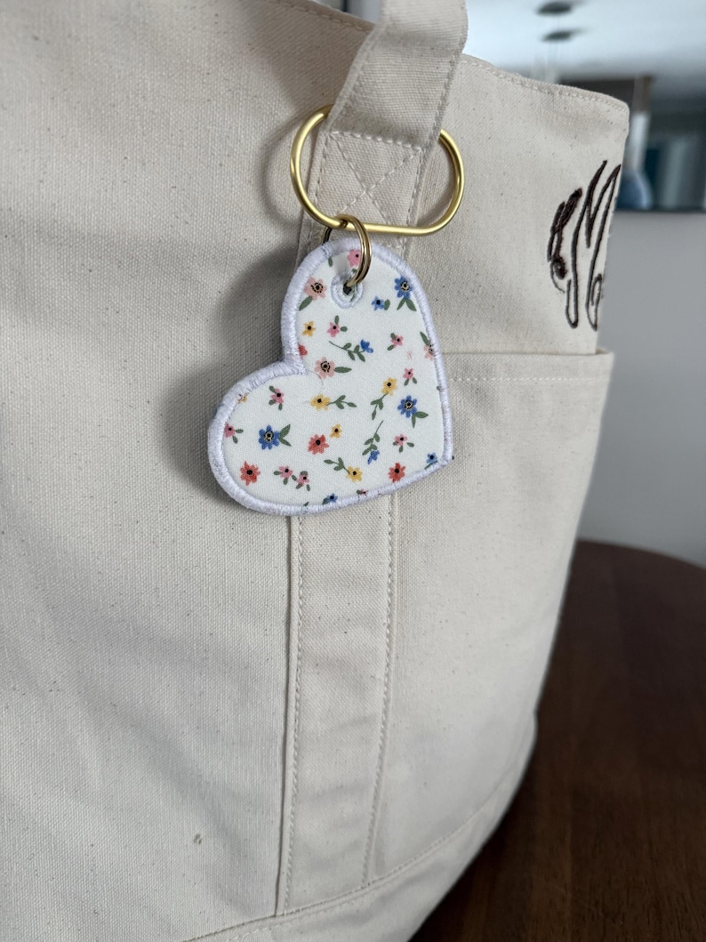 May include: A cream-colored tote bag with a gold-colored ring holding a heart-shaped keychain. The keychain has a white background with a floral pattern in blue, pink, and yellow. The bag has a pocket with embroidered initials.
