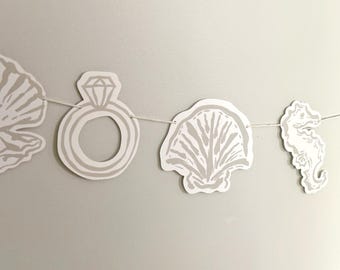 May include: A decorative garland featuring die-cut paper shapes in a light beige color. The shapes include a scallop shell, a diamond ring, another scallop shell, and a seahorse, all strung together on a thin white string for hanging display.
