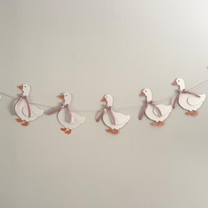 Puede incluir: Una guirnalda decorativa de gansos de papel blanco con patas y picos naranjas, cada uno adornado con una cinta rosa. Los gansos están ensartados en una cuerda blanca, creando una pancarta. Fondo blanco.