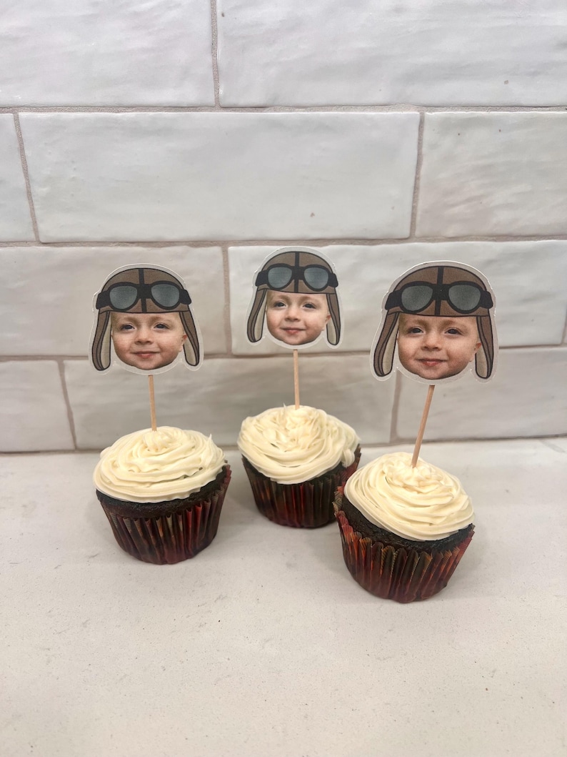 May include: Three chocolate cupcakes with white frosting topped with edible images of a child wearing a brown aviator hat and goggles.