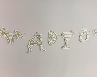 May include: A decorative garland featuring a series of white paper cutouts with sage green line art illustrations. The shapes include a bikini bottom, a bikini top, a citrus slice, a cocktail glass, and a diamond ring, all strung on a thin white cord.