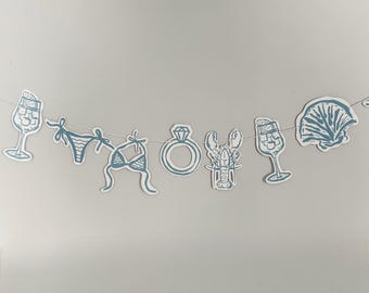 May include: A decorative garland featuring a series of blue and white paper cutouts strung together. The shapes include a cocktail glass, a bikini, a diamond ring, a lobster, another cocktail glass, and a seashell, all in a uniform blue ink style.