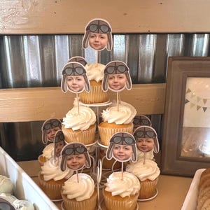 May include: A white cupcake stand with multiple tiers, decorated with cupcakes topped with white frosting and personalised aviator hats. The hats feature a child's face and goggles.
