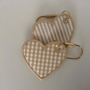 May include: Two heart-shaped keychains. One features a tan and white striped pattern, the other a tan and white gingham pattern. Both have gold-colored metal hardware.