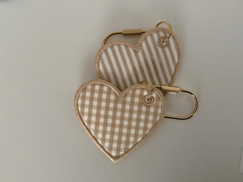 May include: Two heart-shaped keychains. One features a tan and white striped pattern, the other a tan and white gingham pattern. Both have gold-colored metal hardware.