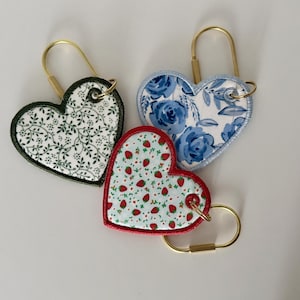 May include: Three heart-shaped keychains with gold-colored hardware. One features a white background with green leaves, another has blue floral patterns, and the third has a red border with a strawberry pattern.