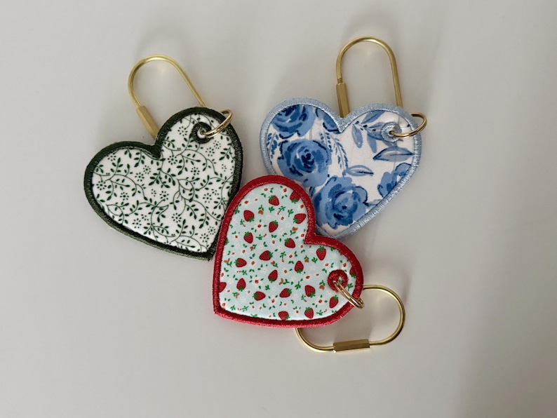 May include: Three heart-shaped keychains with gold-colored hardware. One features a white background with green leaves, another has blue floral patterns, and the third has a red border with a strawberry pattern.