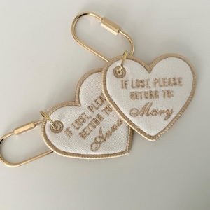 May include: Two heart-shaped keychains with gold-colored hardware. The white fabric hearts have gold embroidered text that reads "IF LOST, PLEASE RETURN TO:" followed by a name. One says "Anna" and the other says "Mary".