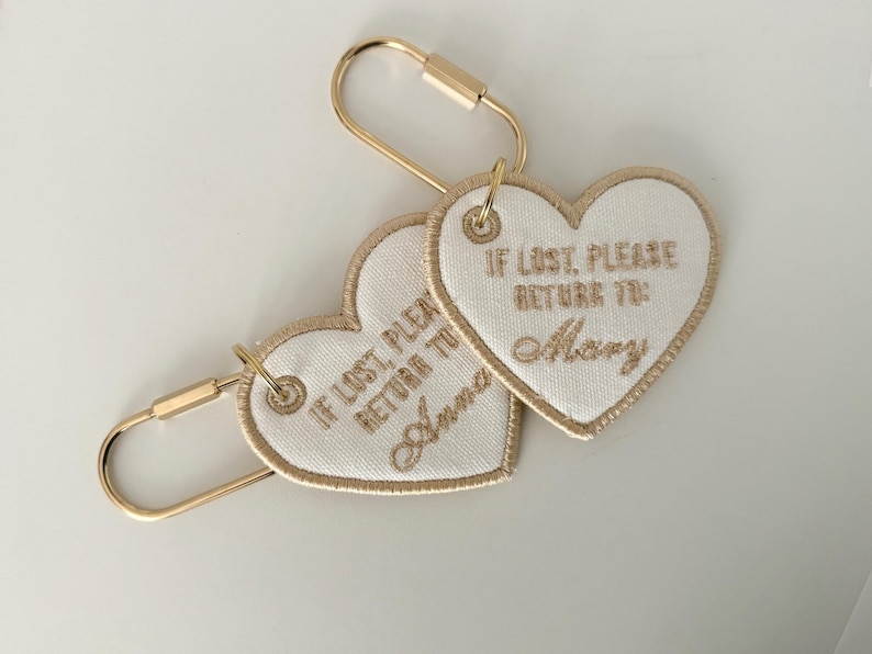 May include: Two heart-shaped keychains with gold-colored hardware. The white fabric hearts have gold embroidered text that reads "IF LOST, PLEASE RETURN TO:" followed by a name. One says "Anna" and the other says "Mary".