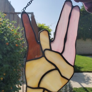 May include: A stained glass window hanging with a chain. The window is shaped like a hand making the 'I love you' sign with two fingers up. The hand is made of yellow, brown, and pink glass.