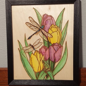 Può includere: Un'opera d'arte pirografata incorniciata con un bouquet di tulipani gialli e viola con due libellule e una rana verde.