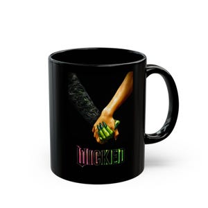 May include: Black ceramic mug featuring a graphic of two hands holding each other, one with green claws and the other with tan skin. The word "WICKED" is printed in a gradient of pink, green, and yellow.