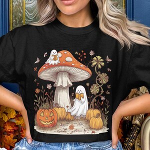 May include: Black t-shirt with a graphic of a mushroom with two ghosts reading books, surrounded by pumpkins and fall foliage.