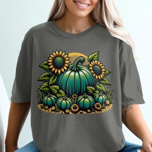 以下が含まれることがあります： 緑の南瓜とひまわりのグラフィックがプリントされたグレーのTシャツ。南瓜は緑の葉と小さな黄色の花に囲まれています。