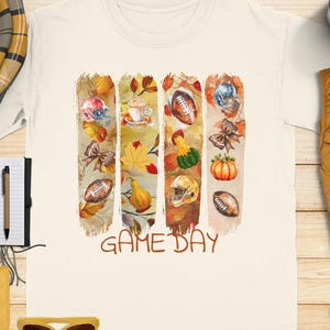 以下が含まれることがあります： 「GAME DAY」のグラフィックが入ったクリーム色のTシャツ。グラフィックは、アメフトボール、ヘルメット、カボチャ、紅葉の垂直な筆致が特徴です。ノート、ペン、サングラスが木製の表面にあります。
